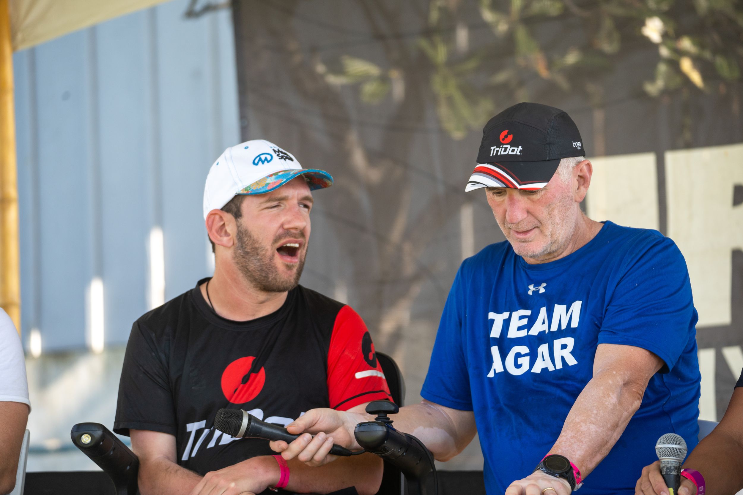 Two people in casual attire share a laugh during an outdoor event, one wearing a black t-shirt and one in blue.