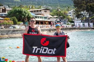 Two people hold up a black and red TriDot banner at a scenic waterfront location with mountains in the background.