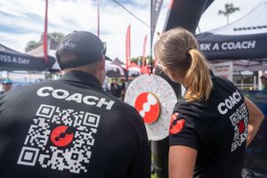 Two coaches wearing black t-shirts with QR codes stand at a sports event with red and white banners visible.