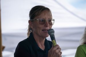 Someone speaks into a microphone while wearing glasses and a dark shirt under a white tent canopy.