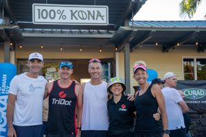 A group of people wearing casual attire and hats stand together outside a storefront with a '100% KONA' sign.