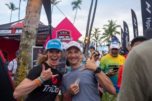 Athletes celebrate and give thumbs up at a triathlon event with palm trees and sponsor banners in the background.