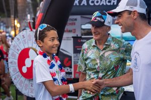 People greet each other at an outdoor event wearing casual attire including a Hawaiian shirt and trucker hat.