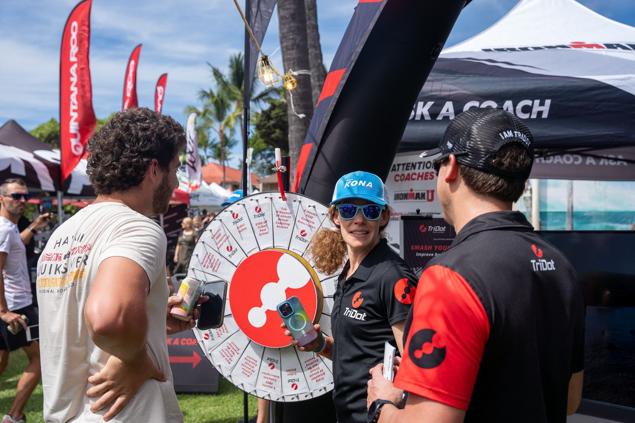 Athletes and event staff gather around a large prize wheel display at an outdoor sports competition with red banners and palm trees.