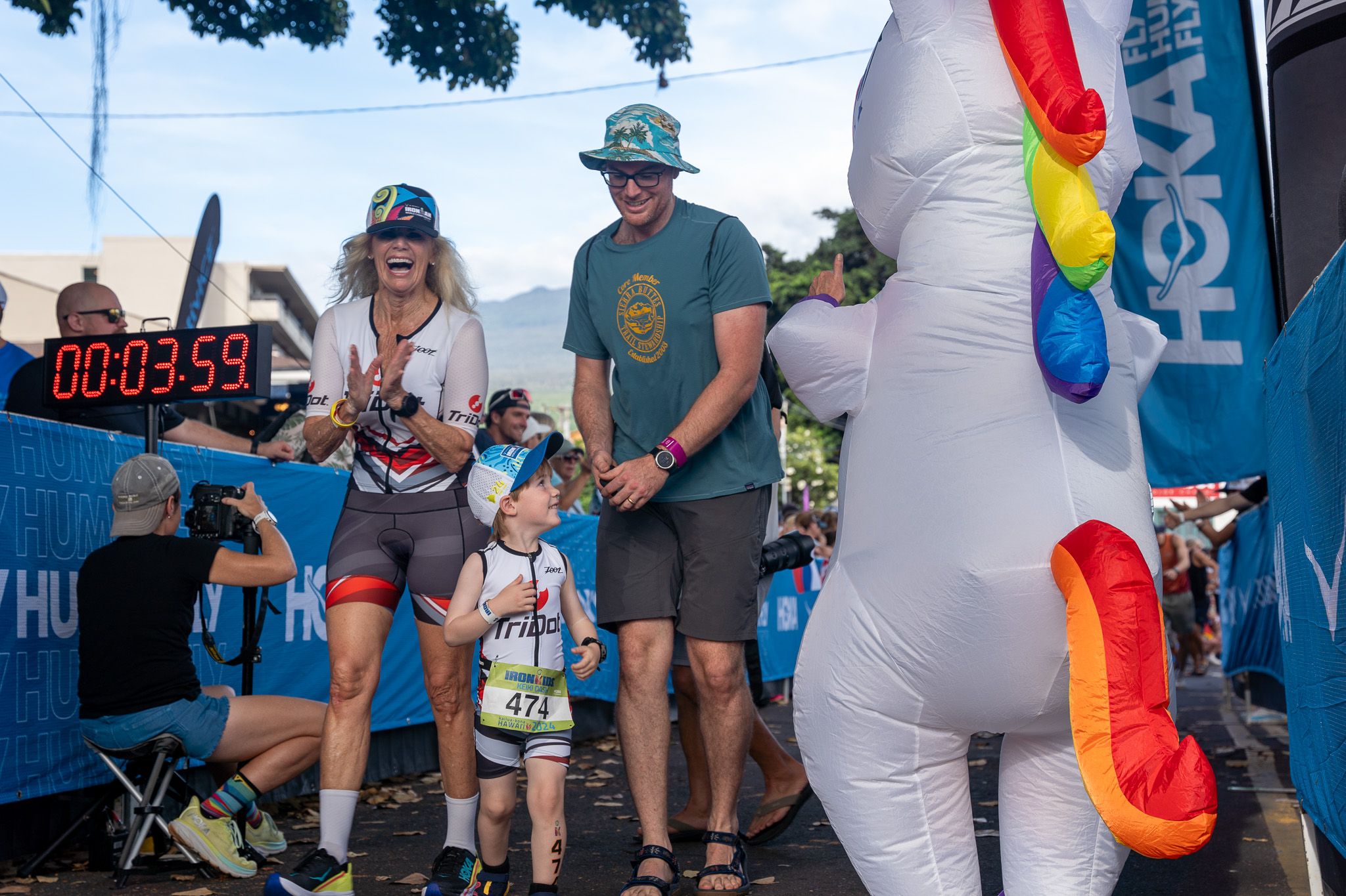 Spectators and supporters gather at a race finish line with an inflatable unicorn decoration and timing clock.
