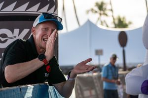 Person in black shirt and blue cap gestures excitedly while standing outdoors with palm trees and tent in background.