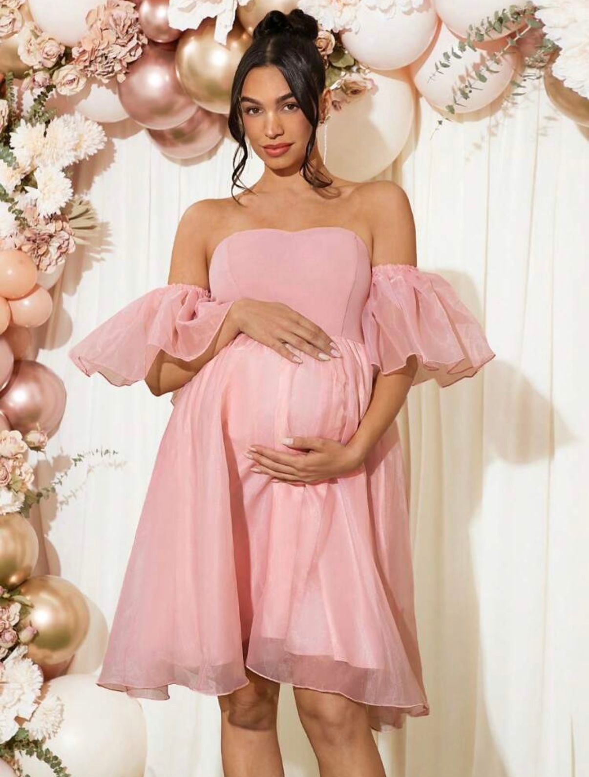 Pink off-shoulder maternity dress with puff sleeves against balloon and floral backdrop.