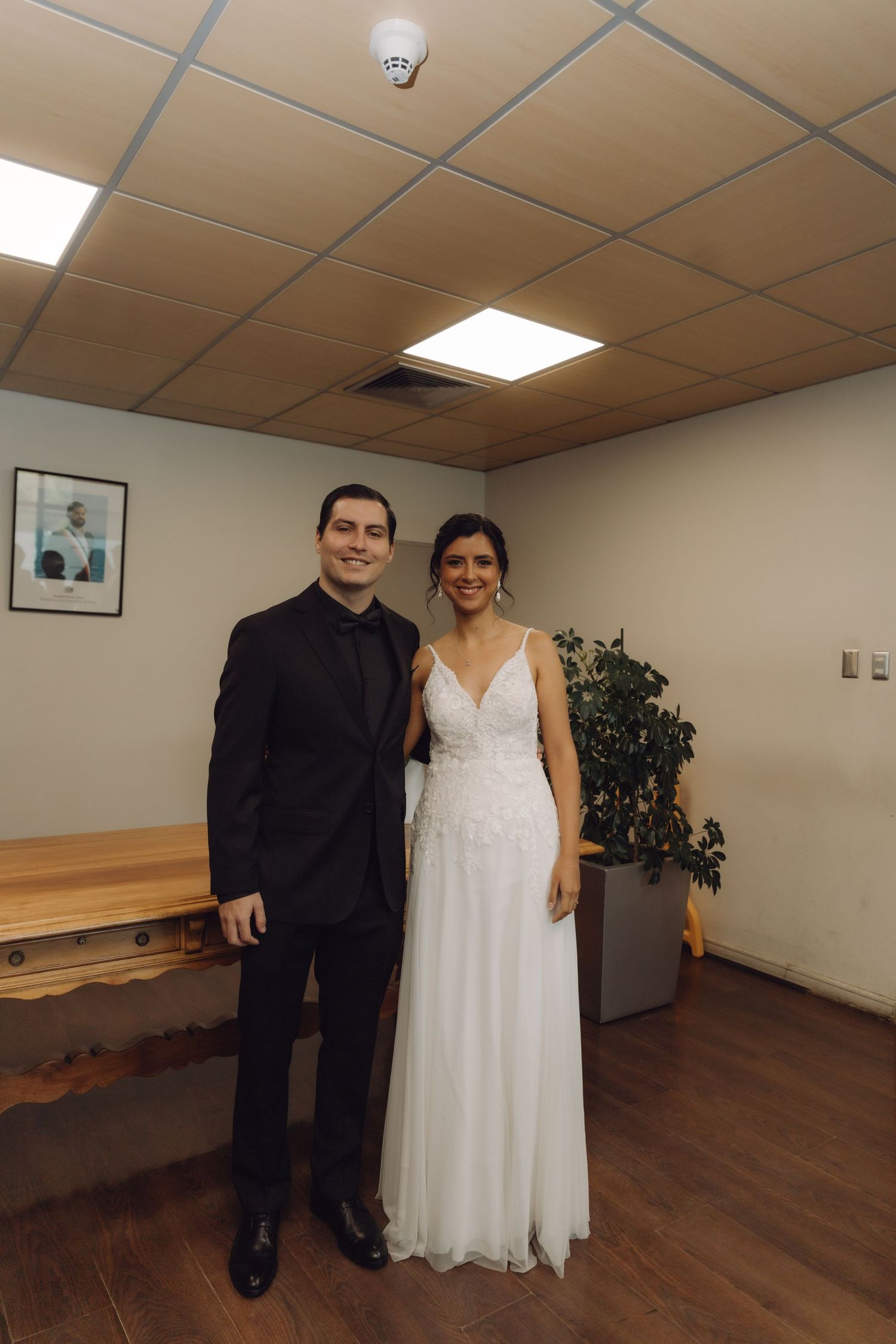 Bride in white gown and groom in black suit posing together in an indoor civil ceremony room.
