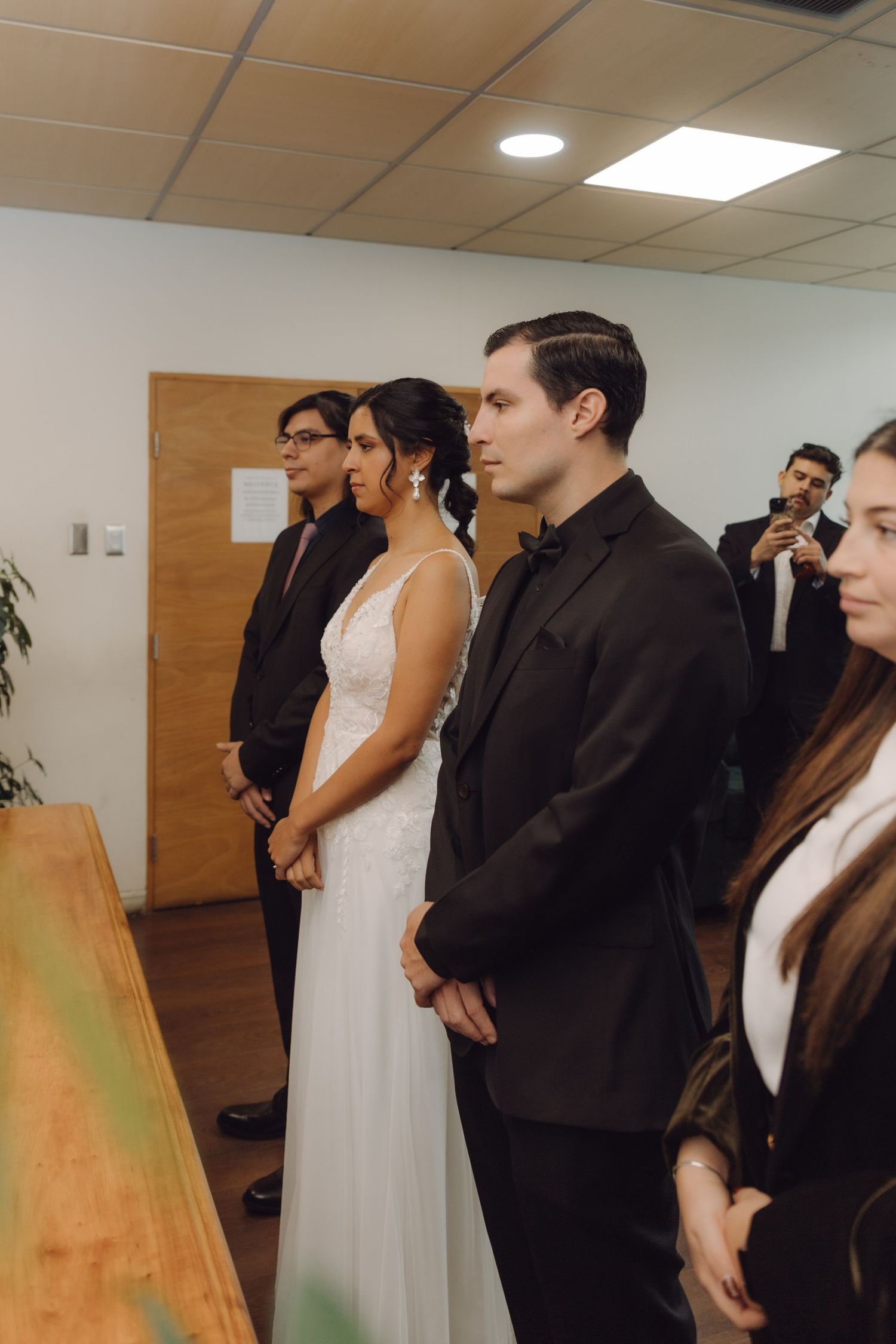 Bride in white dress and groom in black suit standing at a civil wedding ceremony indoors.
