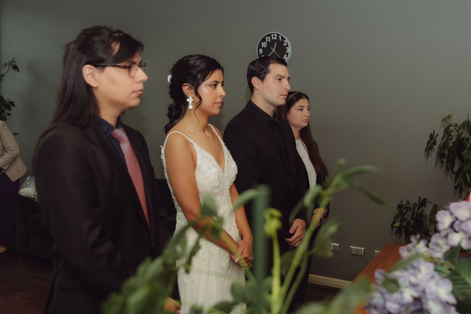 Bride in white gown standing with wedding party guests during an indoor civil marriage ceremony.