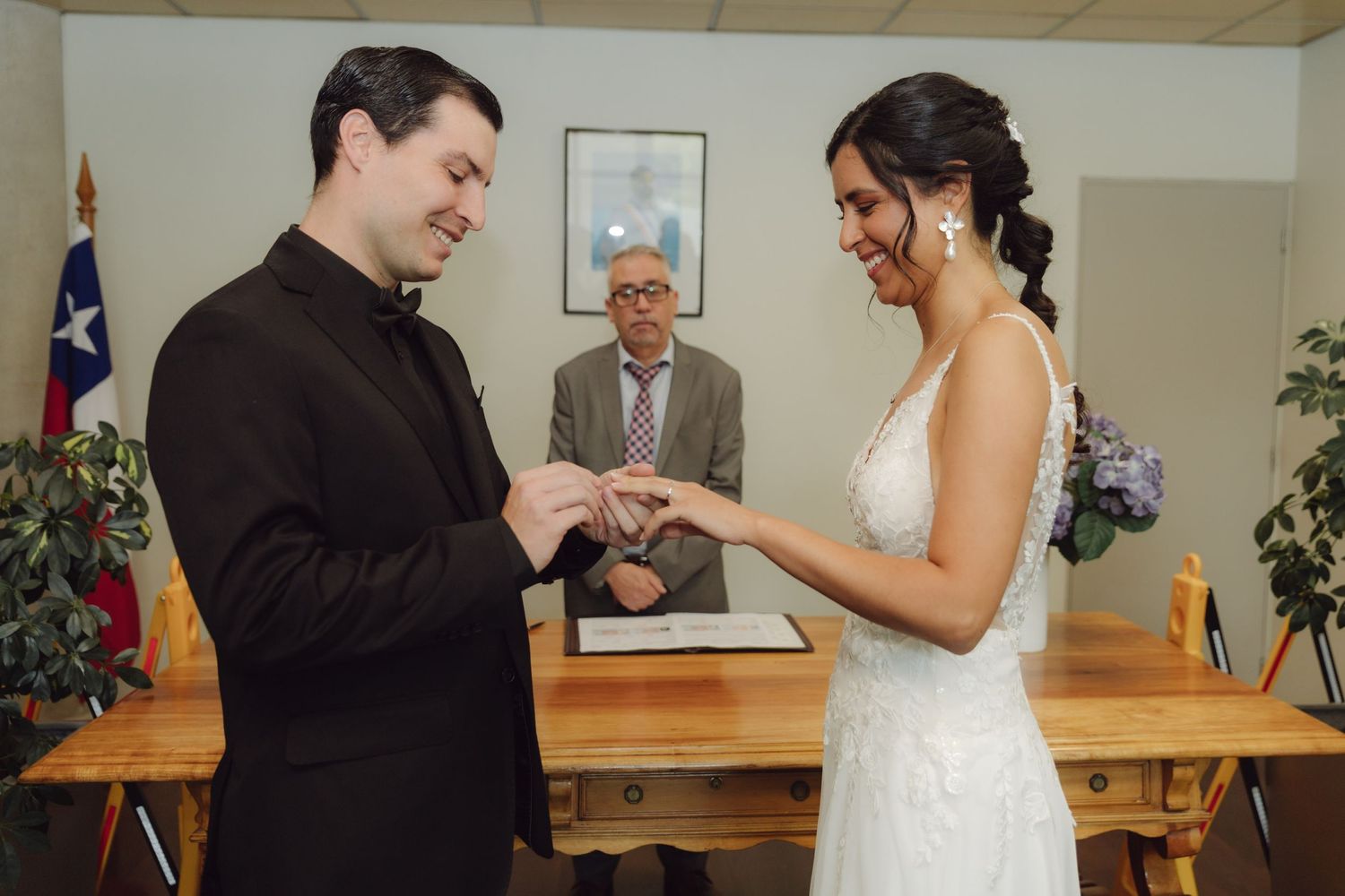 Groom placing wedding ring on bride's finger during civil ceremony with officiant in background.