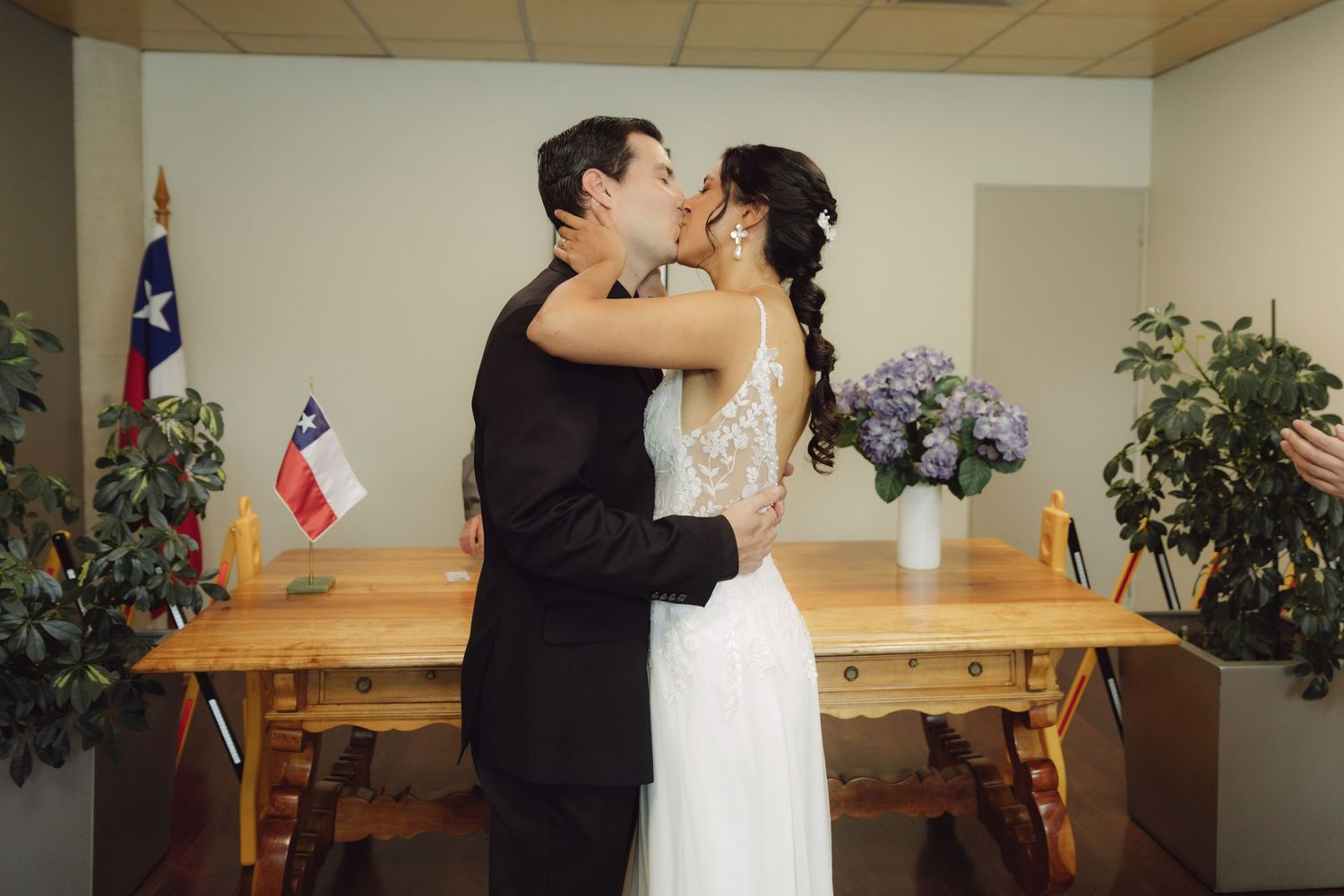 Newlywed couple shares first kiss beside Chilean flag at civil wedding ceremony.