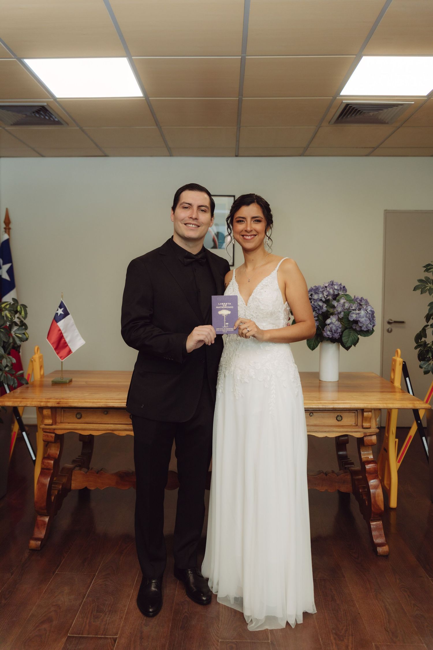 Smiling newlyweds pose with marriage certificate beside Chilean flag after civil ceremony.