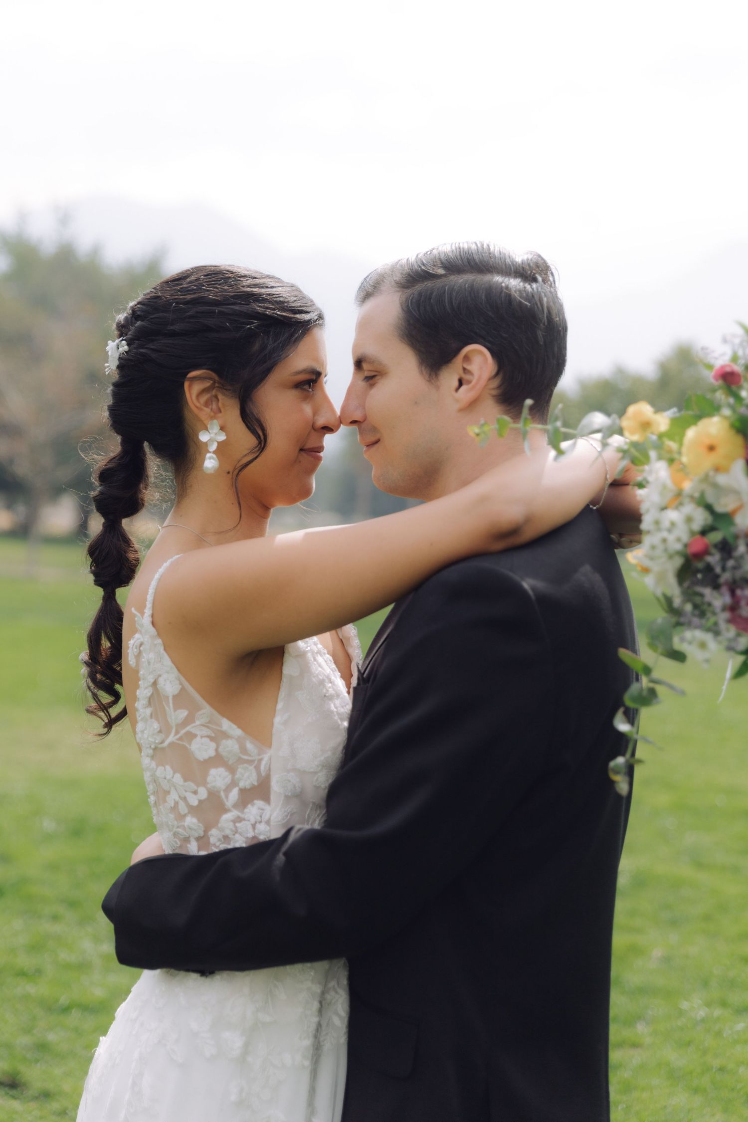 Bride and groom embracing outdoors, nearly kissing while holding a colorful wedding bouquet.