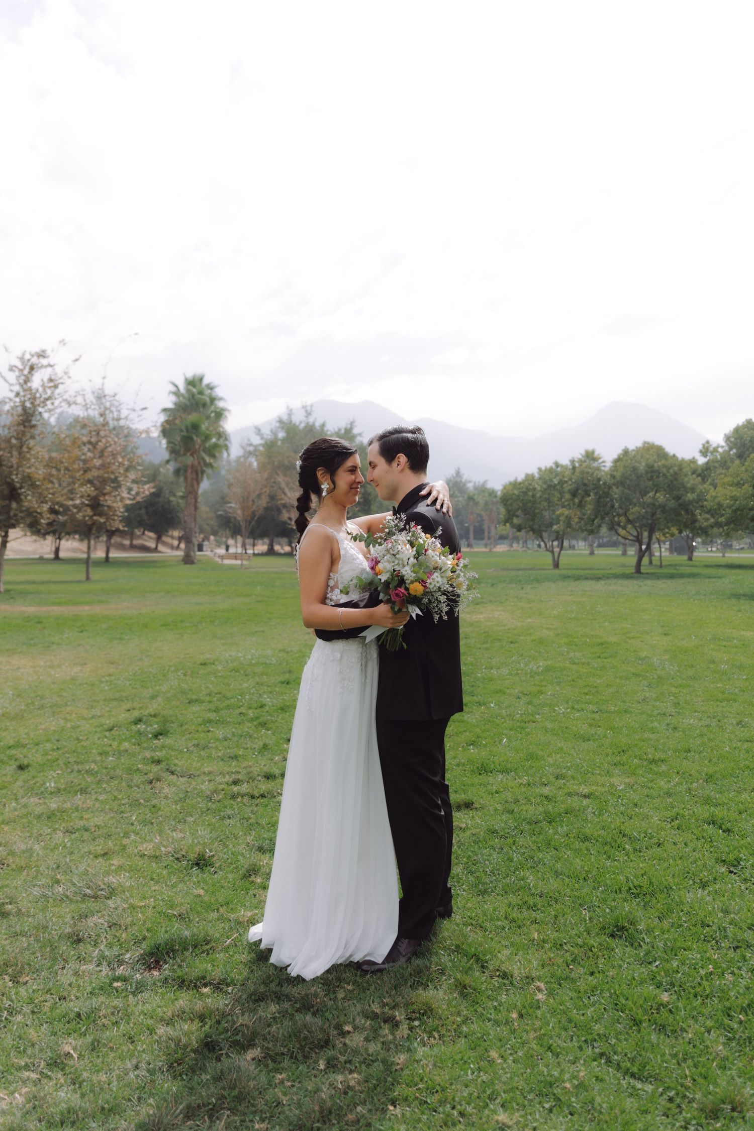 Bride and groom holding bouquet while embracing in a lush green park with mountain views.
