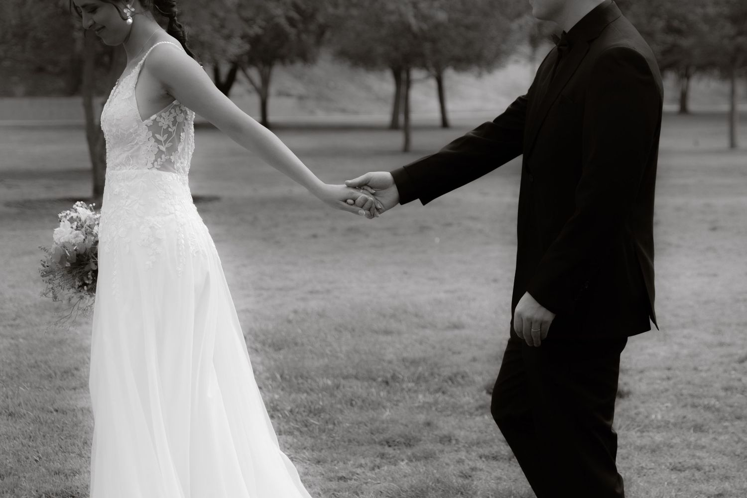 Black and white photo of bride and groom holding hands while walking across a grassy field.