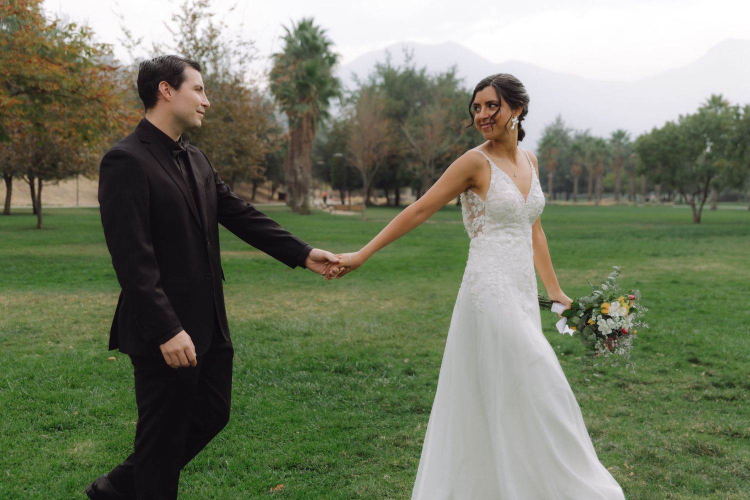 Bride in lace gown and groom in black suit holding hands while walking through a green park.