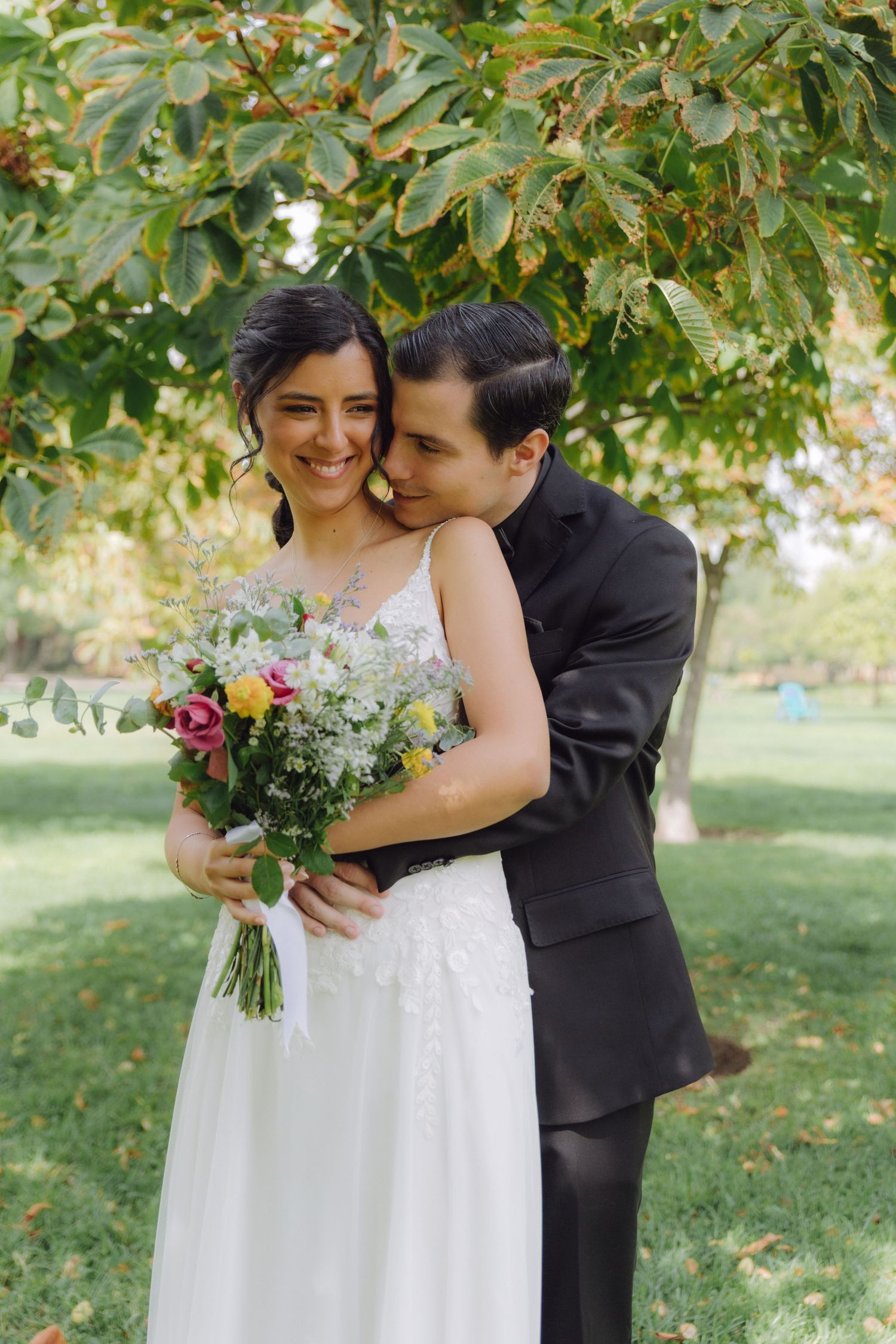 Groom embracing bride from behind under a leafy tree while she holds a colorful bridal bouquet.