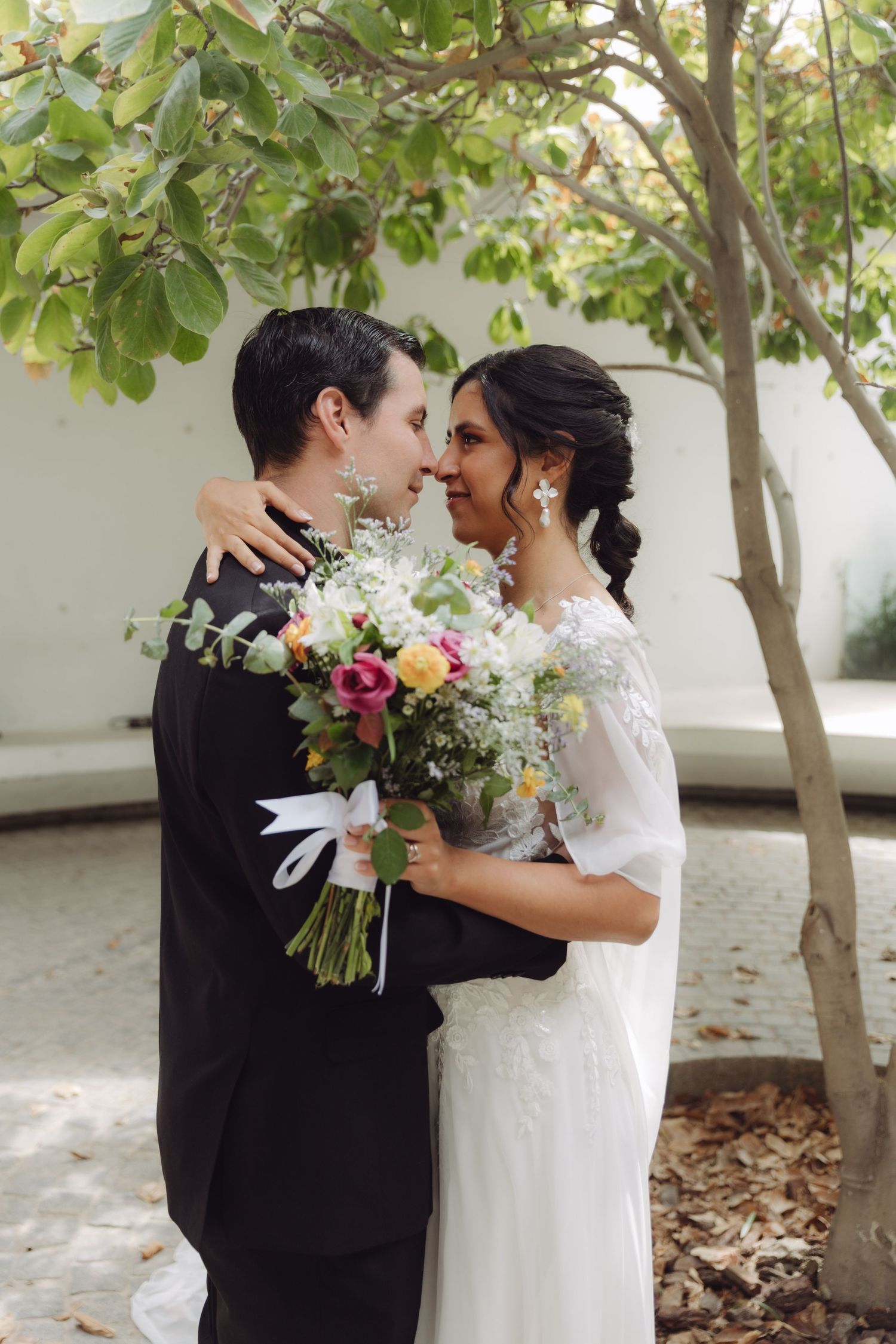 Bride and groom nearly kissing while embracing and holding a colorful bouquet near a tree.