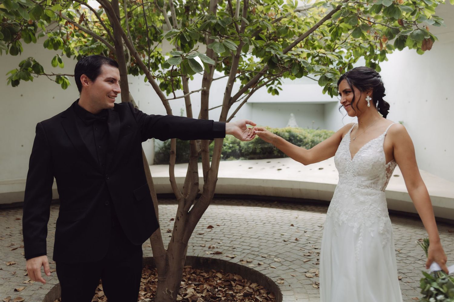 Bride and groom playfully reaching for each other near a tree in a sunlit courtyard.