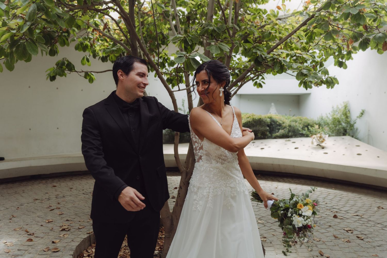 Bride in white lace gown and groom in black suit laughing together outdoors near a tree.