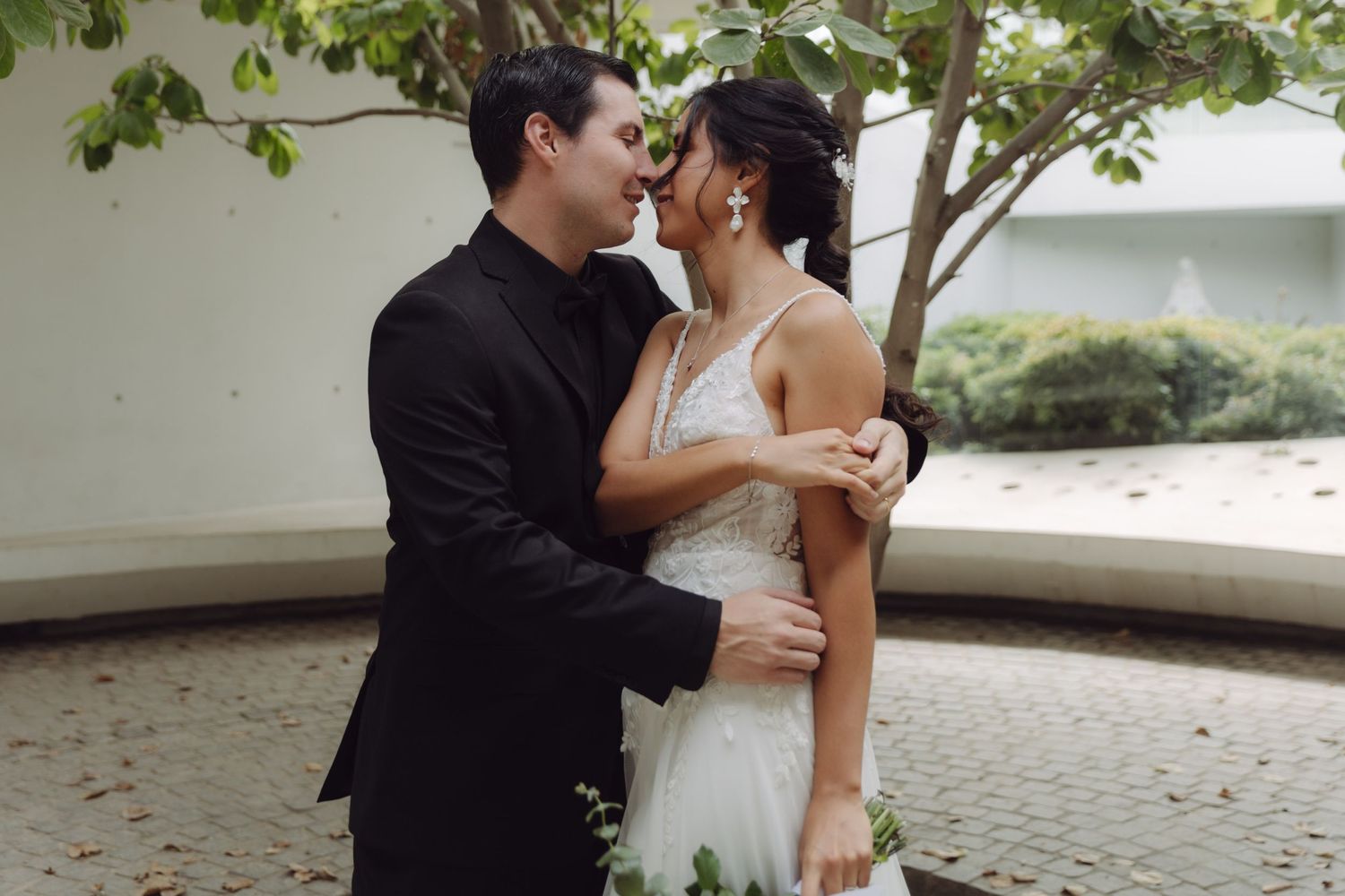 Bride and groom sharing a romantic moment, nearly kissing, embracing under a tree outdoors.