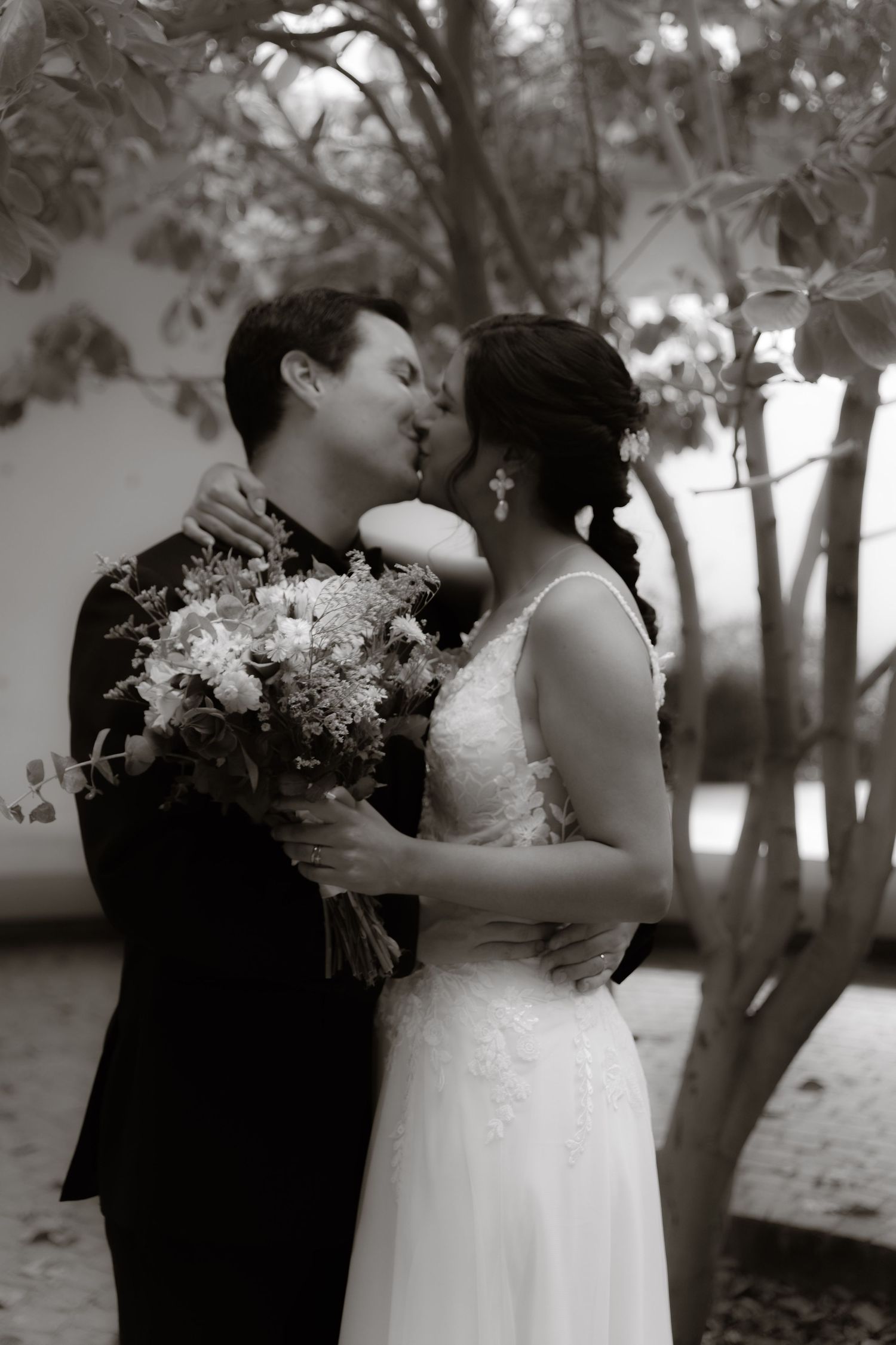 Black and white photo of bride and groom kissing while holding a floral bouquet under trees.