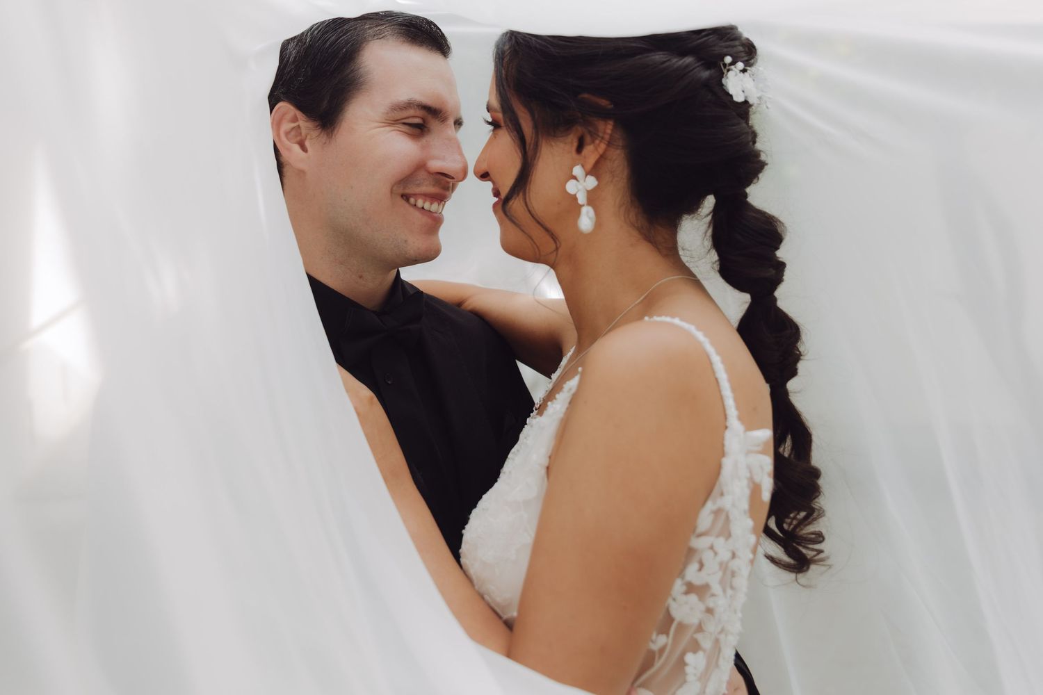 Bride and groom smiling at each other closely, partially wrapped in a flowing white veil.