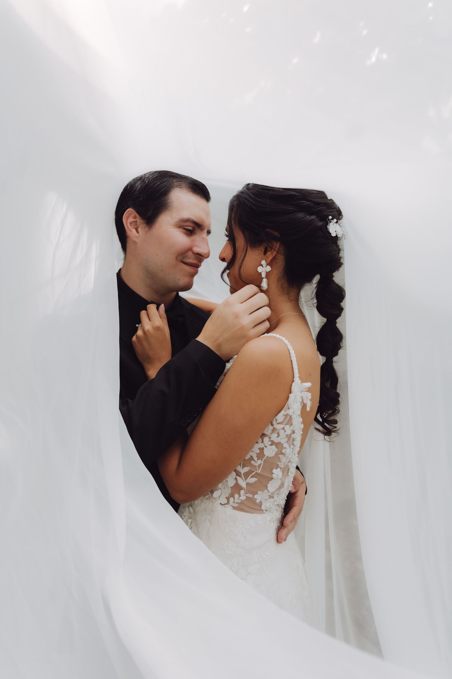 Groom tenderly touching brides face as they gaze at each other surrounded by white fabric.