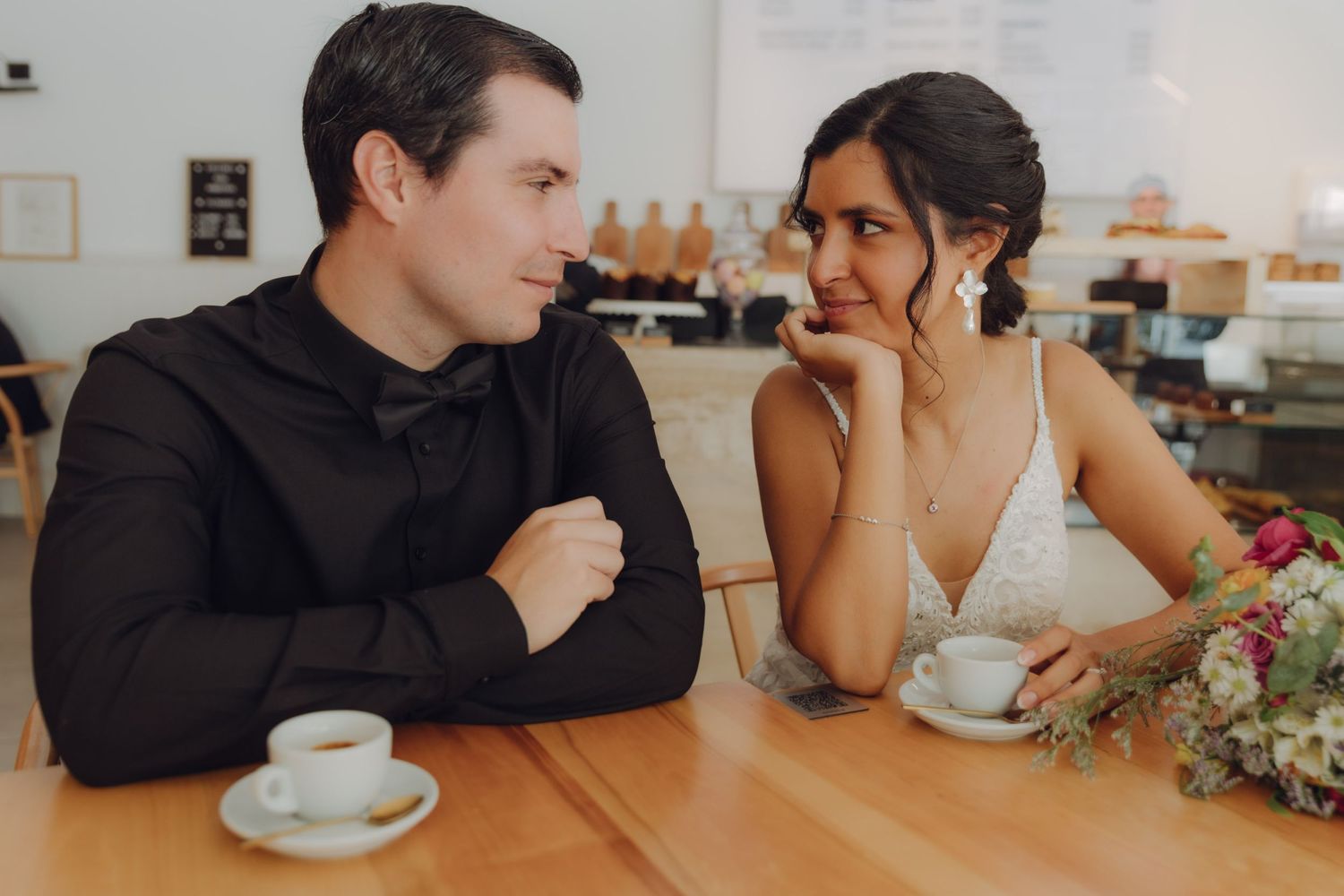 Bride and groom sitting at a cafe table with espresso cups, smiling at each other warmly.
