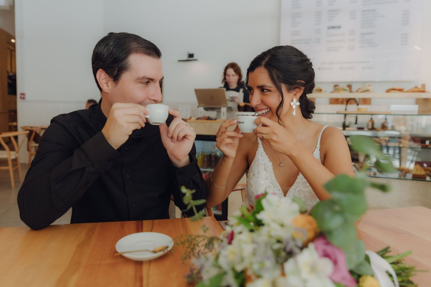 Smiling bride and groom sip espresso together at a cafe on their wedding day.