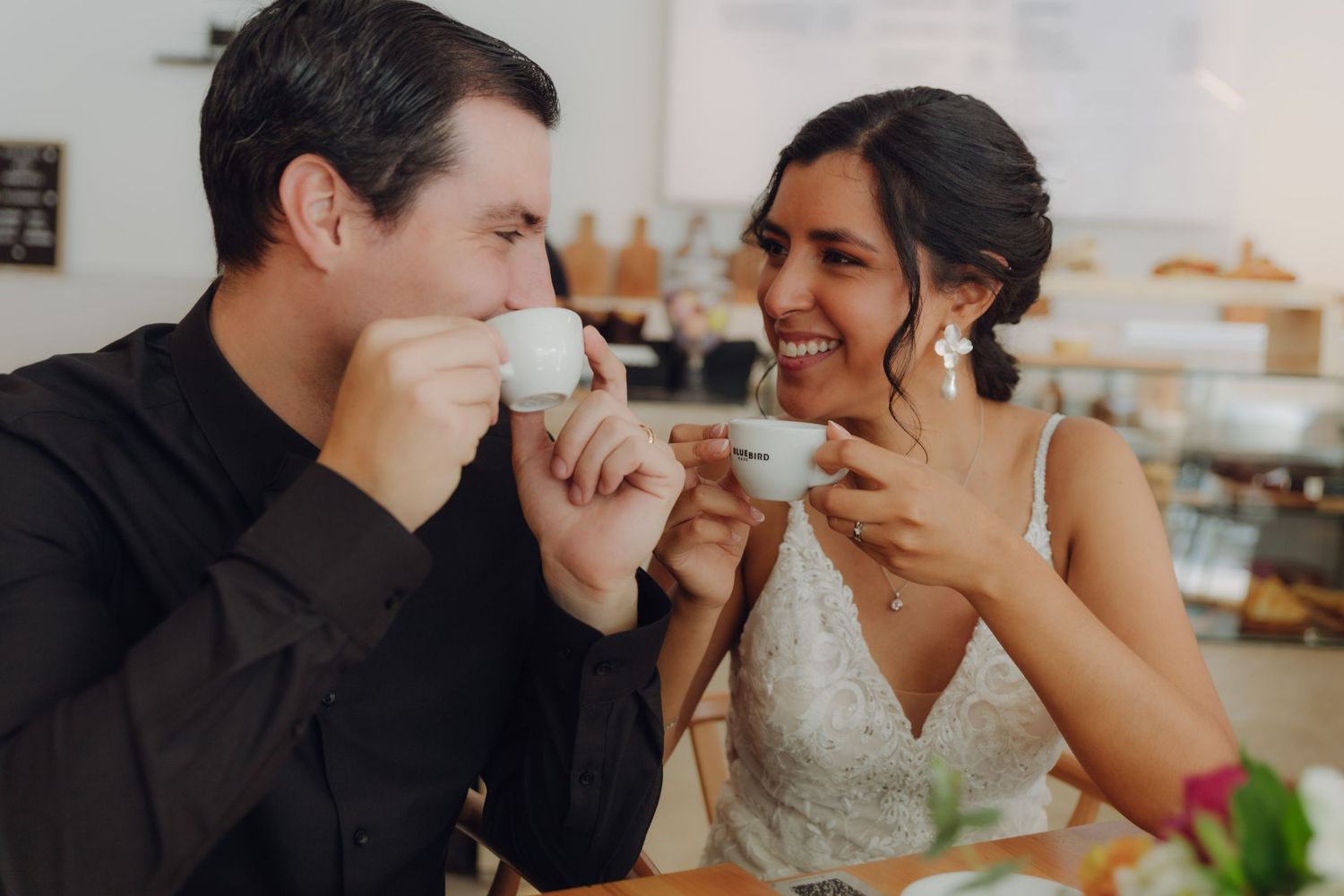 Happy newlyweds clink espresso cups and laugh together at a cafe table.