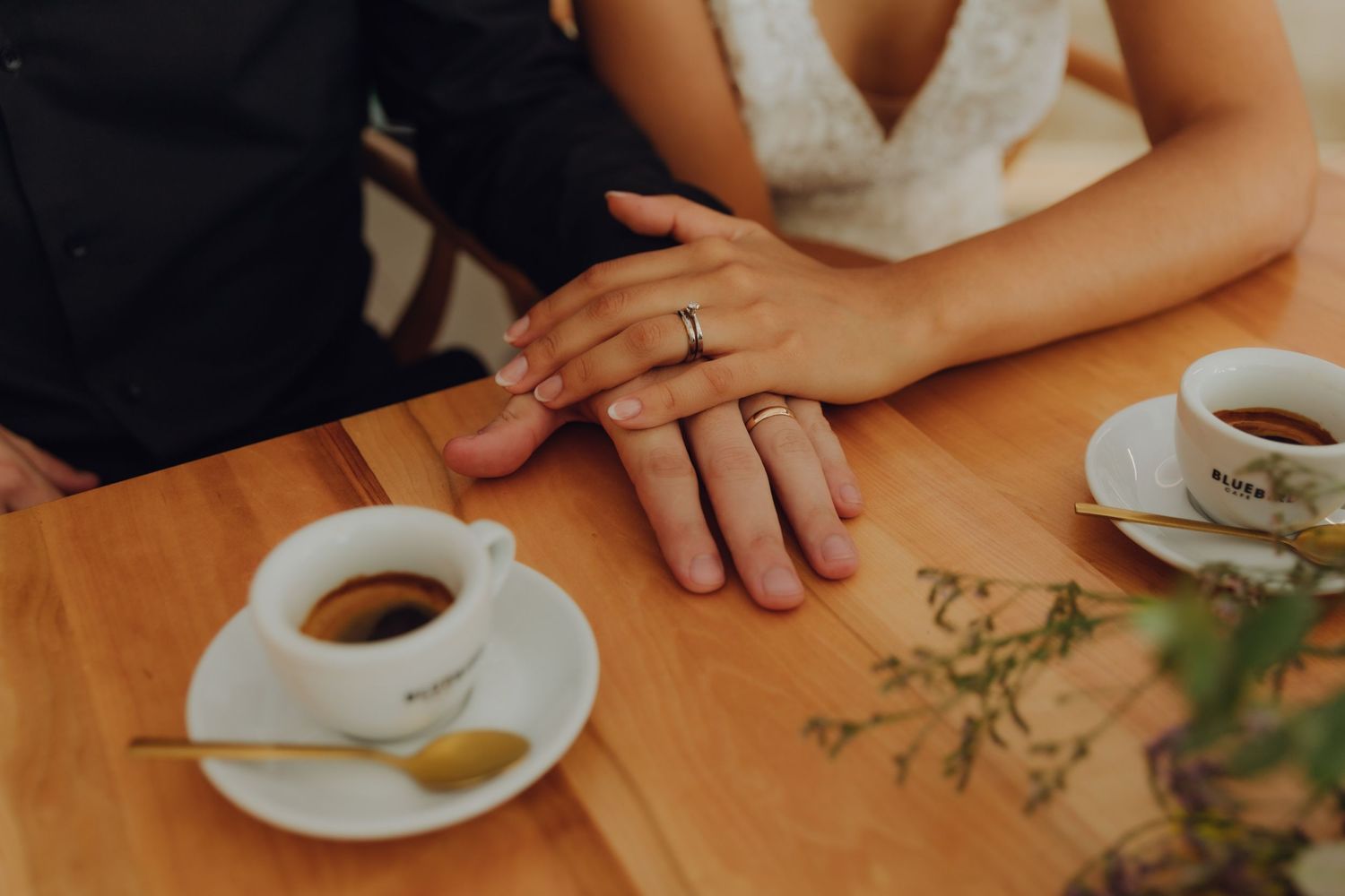 Newlyweds display wedding rings with hands resting near espresso cups on table.