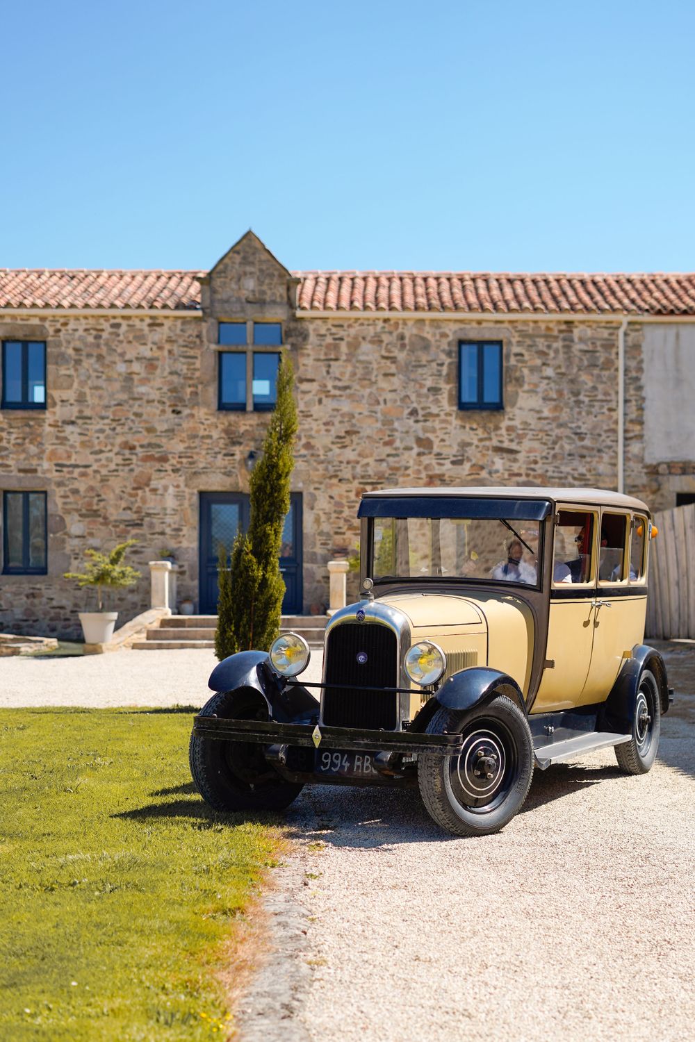 Vintage yellow car parked in front of rustic stone building with blue windows.