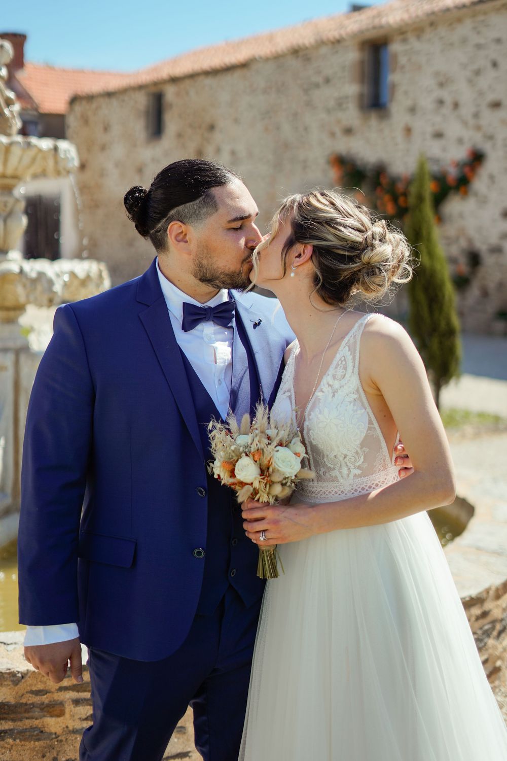 A romantic wedding couple sharing a kiss in front of a rustic stone building with the bride holding a bouquet.