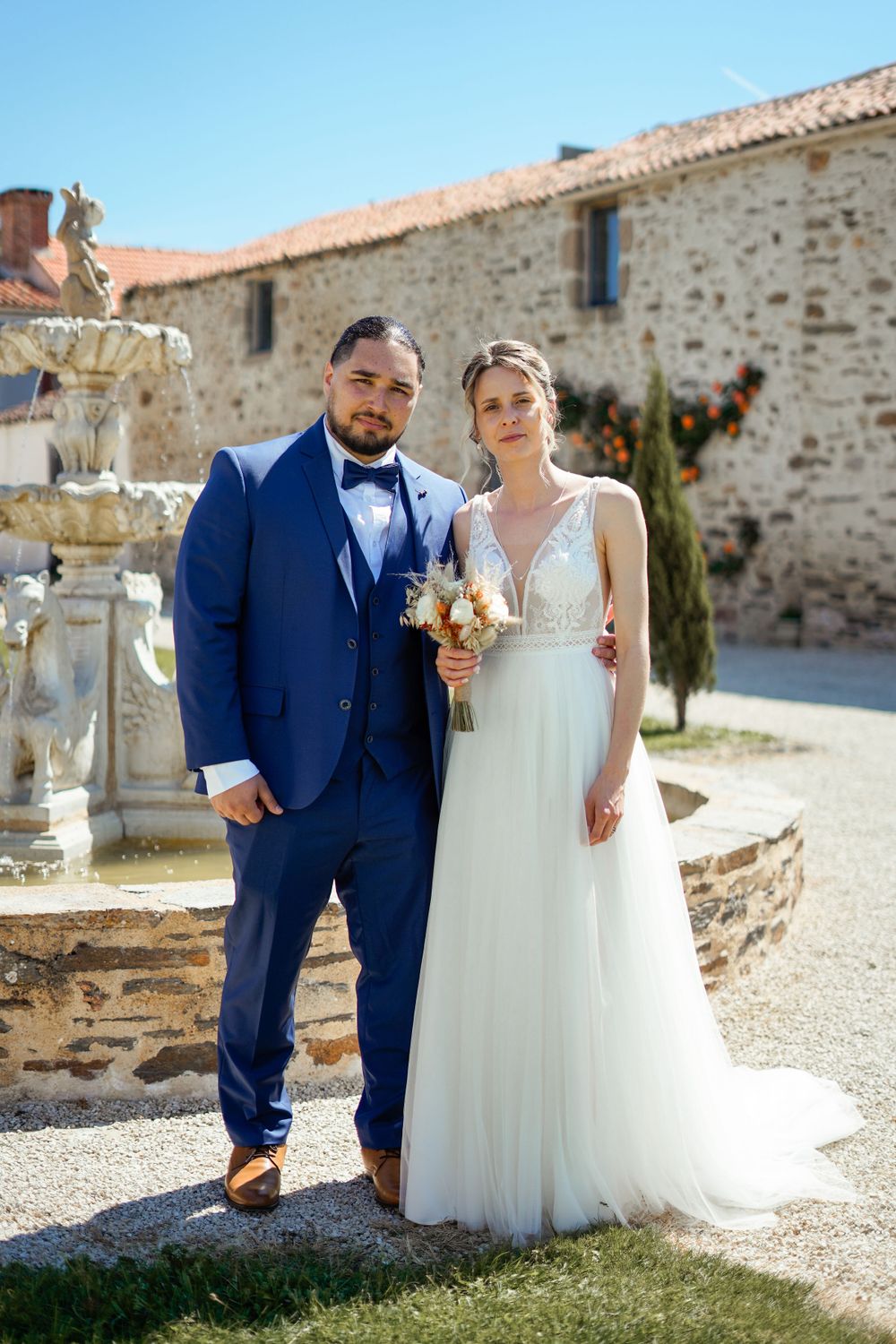 A newlywed couple in blue suit and white dress pose together by a stone wall and fountain.