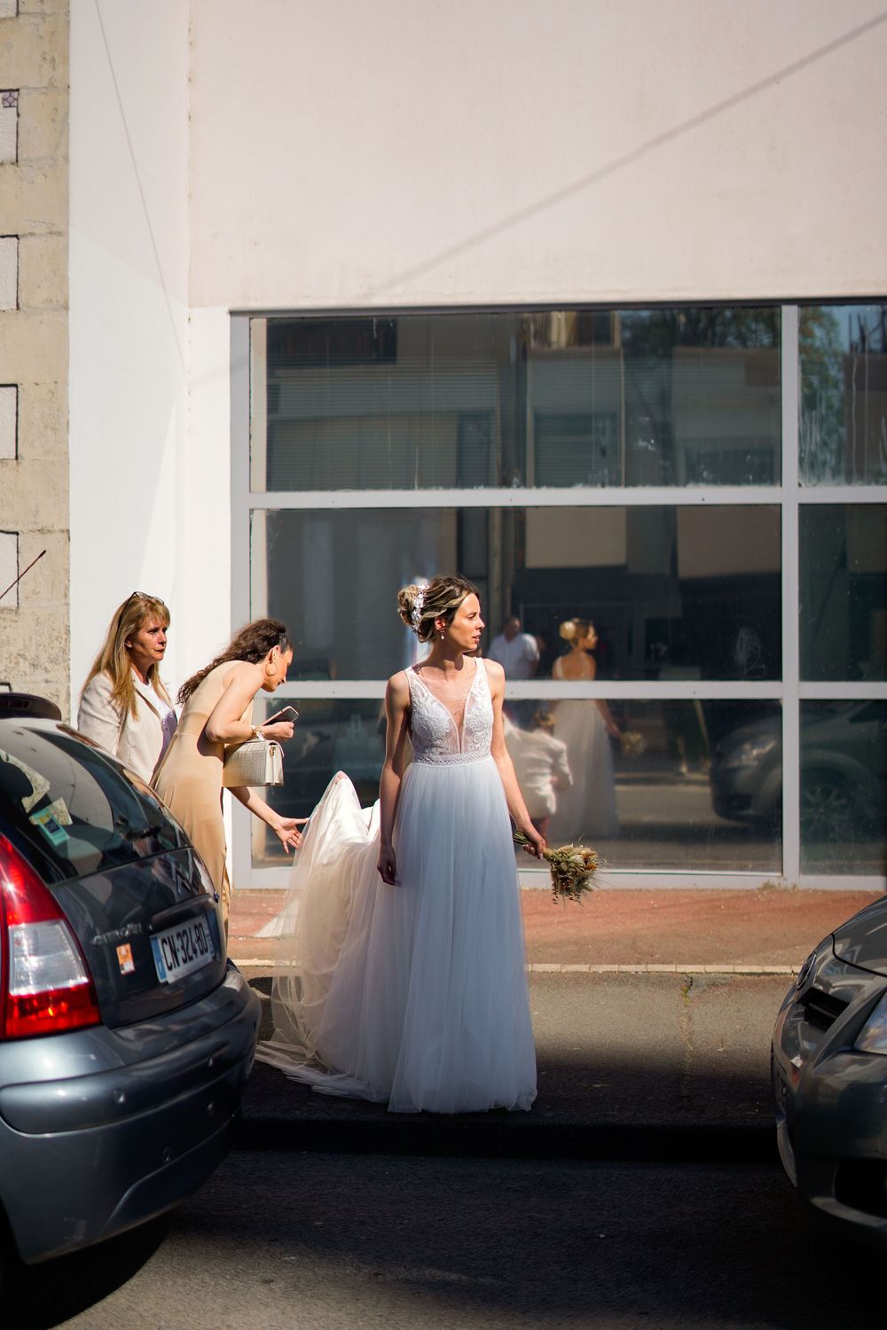 Bride in flowing white dress standing between parked cars outside a modern building.