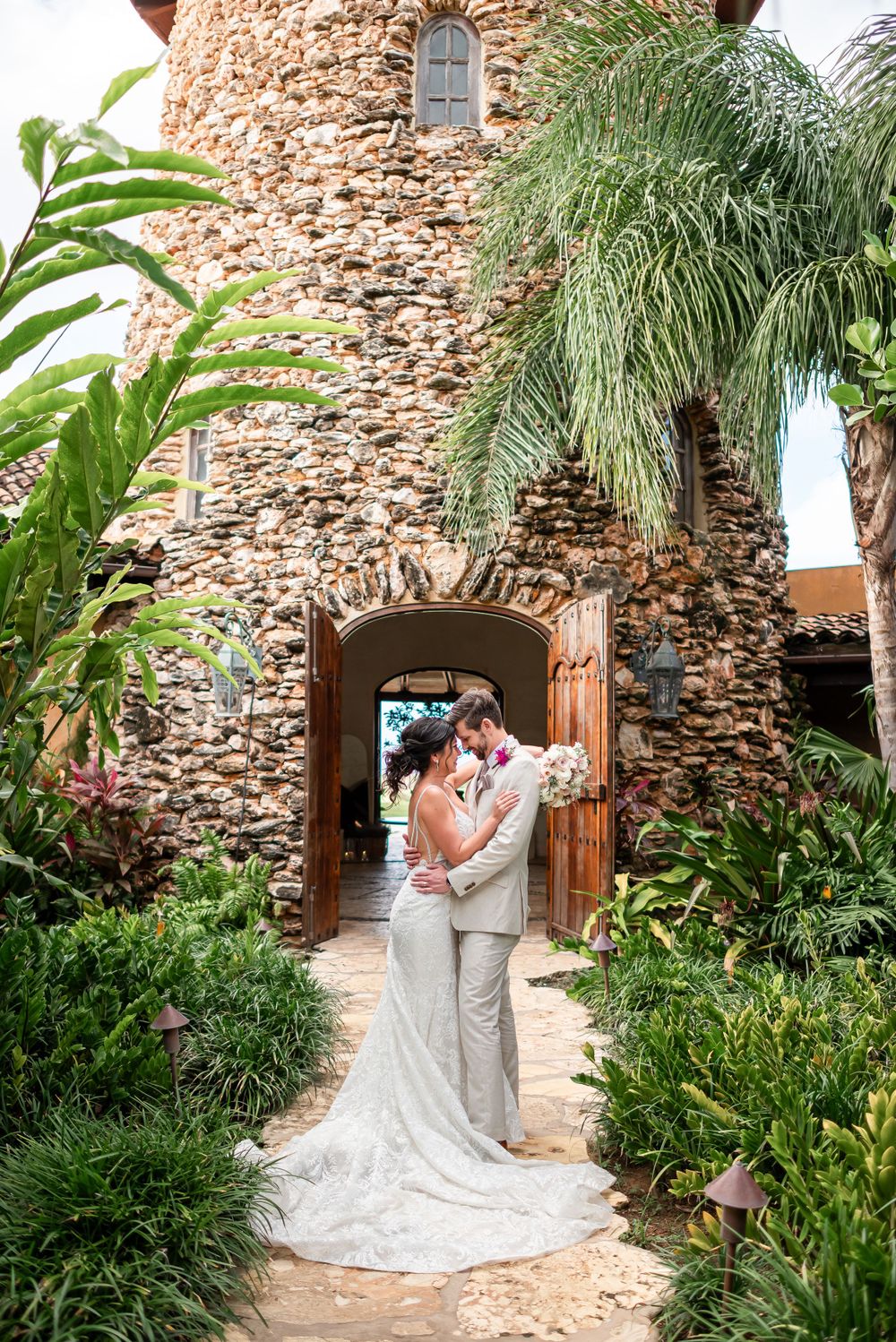 Stone tower entrance framed by tropical plants creates dramatic backdrop for wedding portraits.