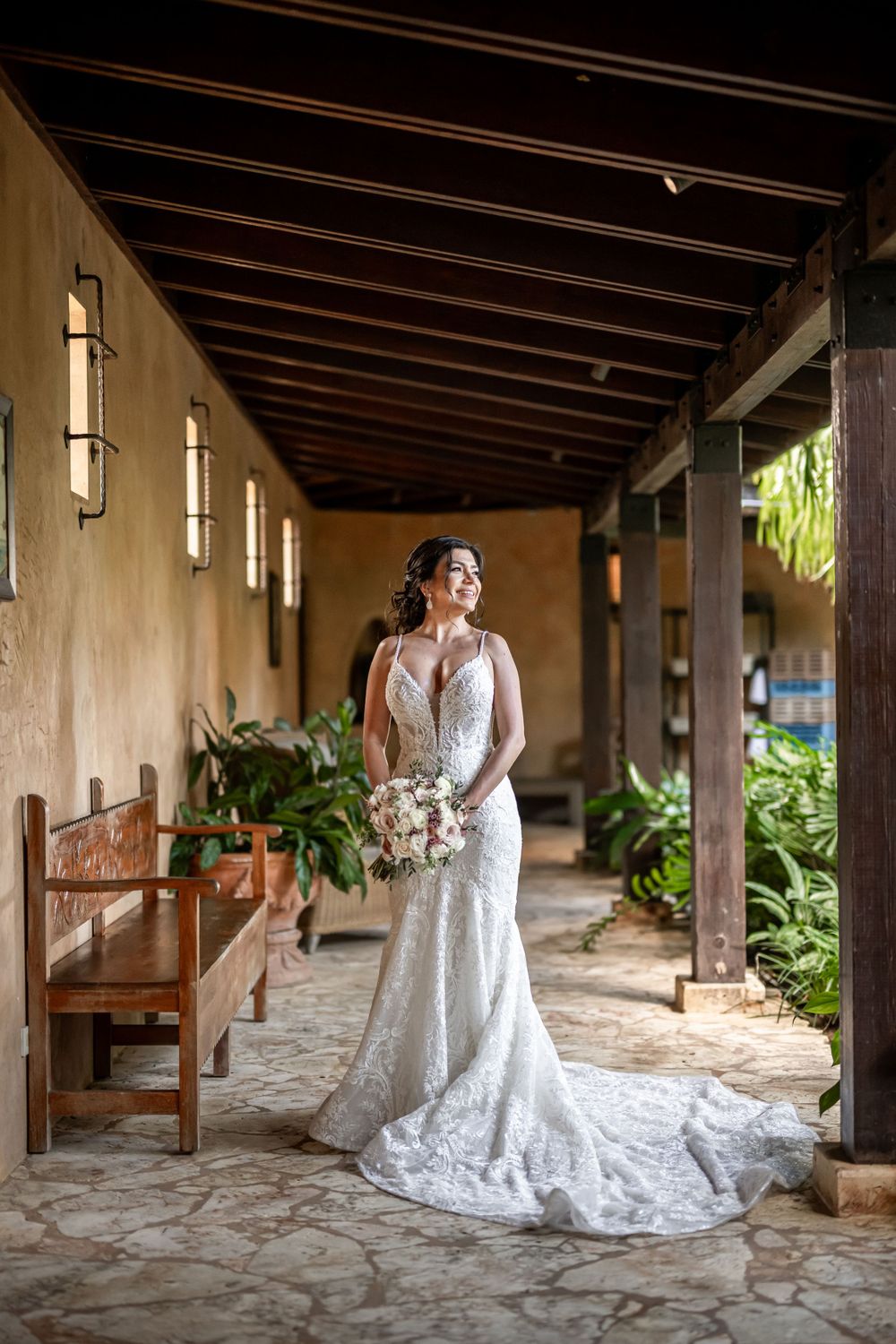 Elegant bride in lace wedding dress poses in Spanish colonial corridor with stone floors and wooden beams.