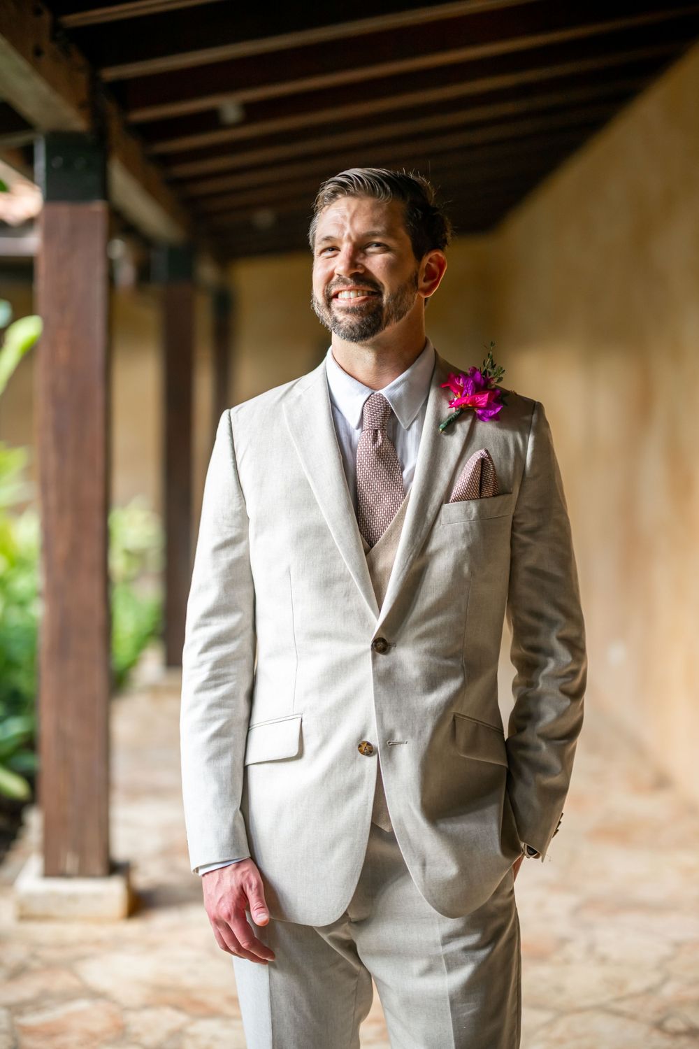 Groom in light beige suit with pink boutonniere stands in covered outdoor walkway.
