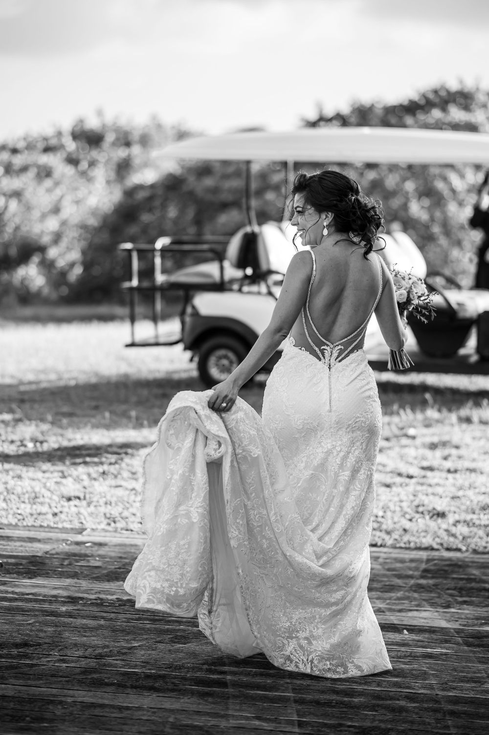 Bride in backless lace gown holds dress train in black and white photo.