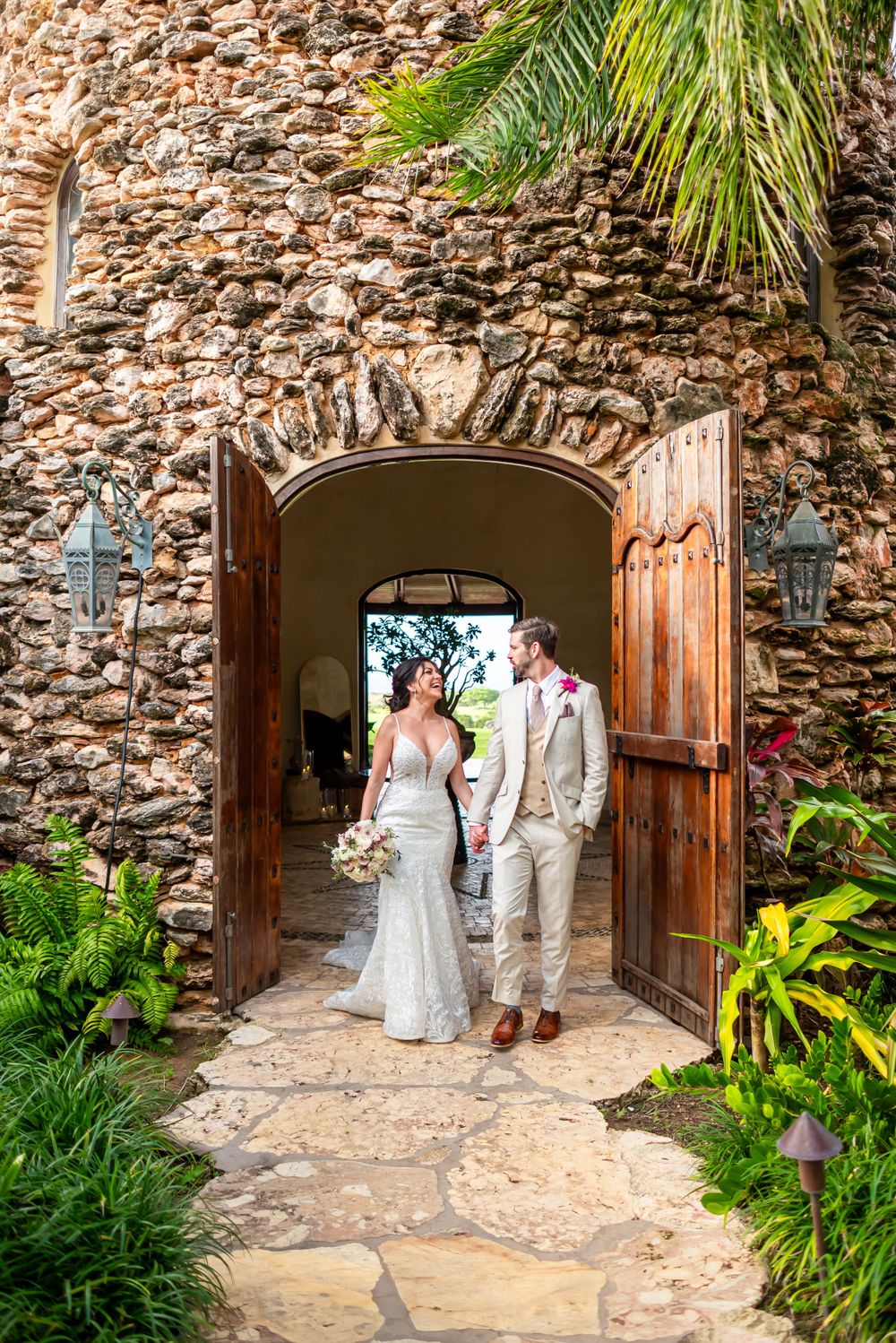 Wedding couple emerging through large wooden doors of a rustic stone building with tropical landscaping.