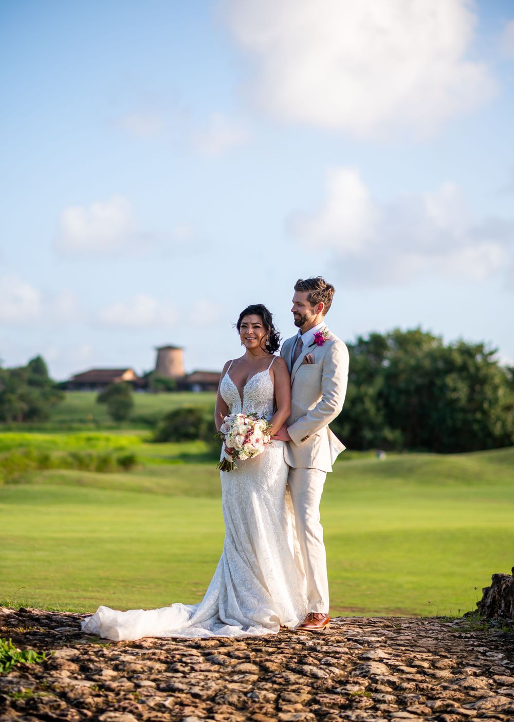 Wedding couple poses on stone wall with green golf course and buildings in background.