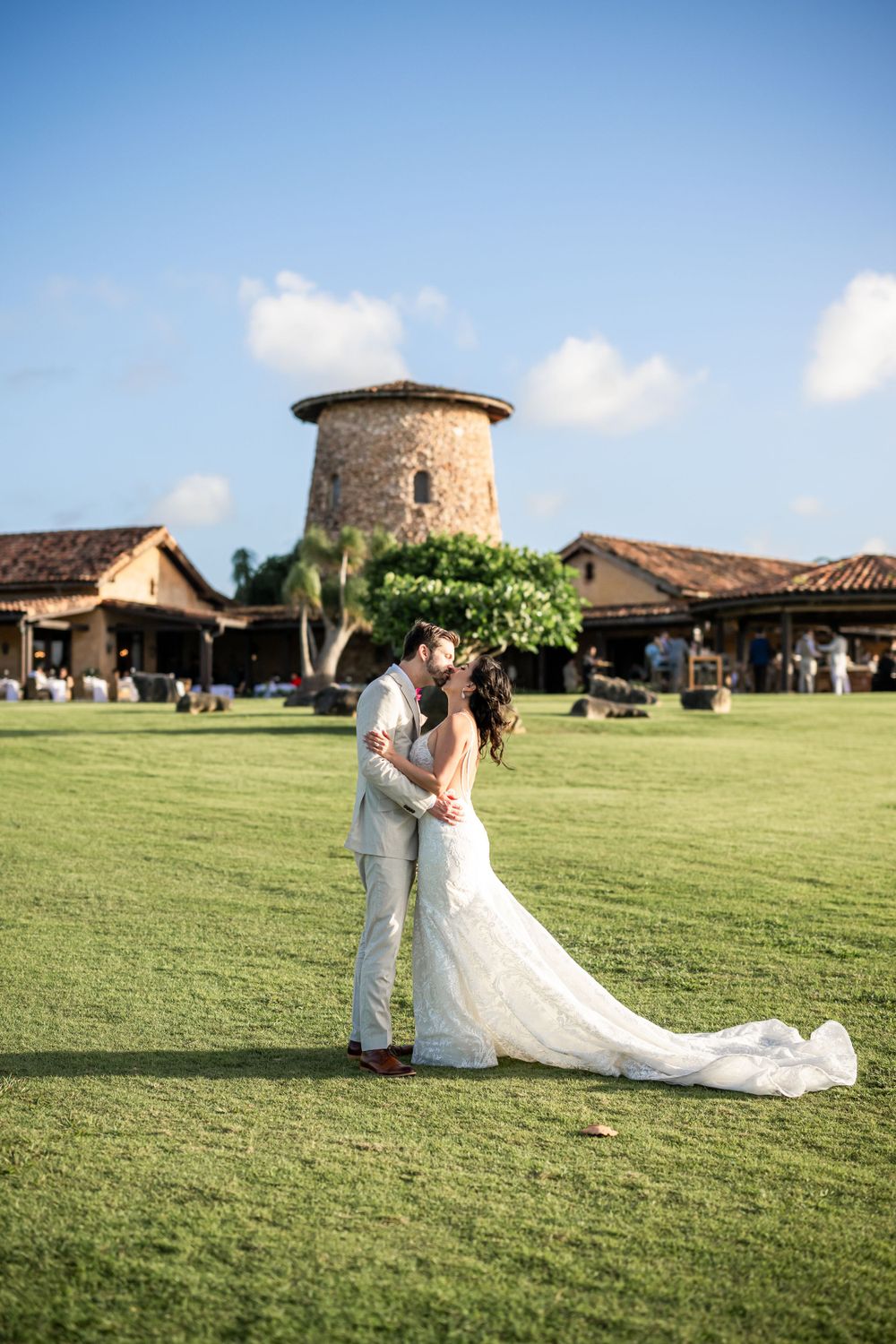 A romantic wedding photo on a lush lawn with a Spanish-style stone tower and terracotta roofs in the background.