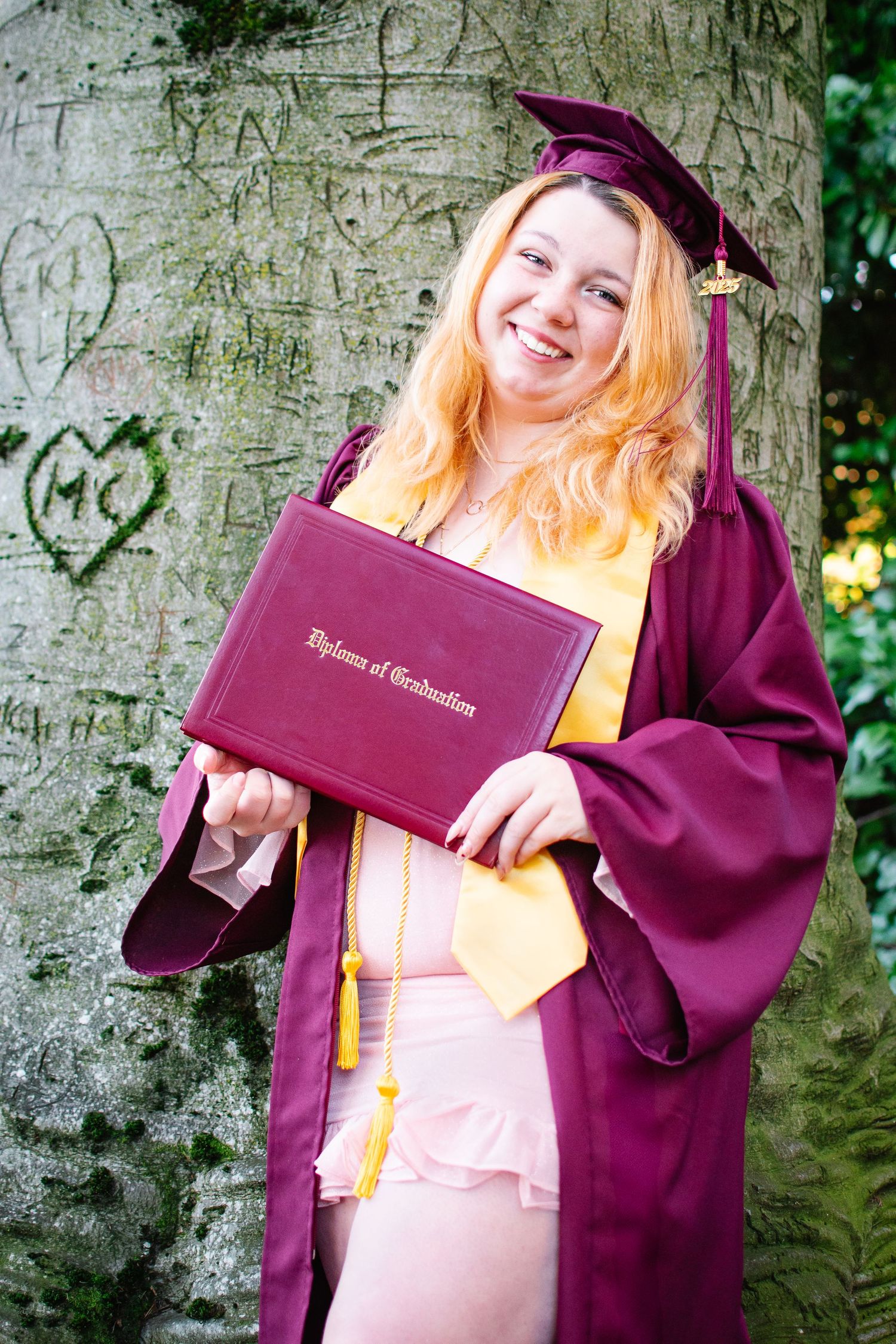 Full-body senior photo of graduate in cap and gown proudly holding diploma in front of tree at Kerry Park in Seattle.