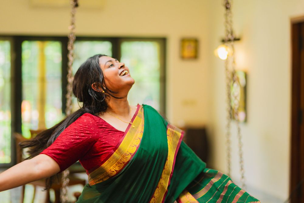 A person in a red and green saree spins joyfully with arms outstretched in a sunlit indoor setting.