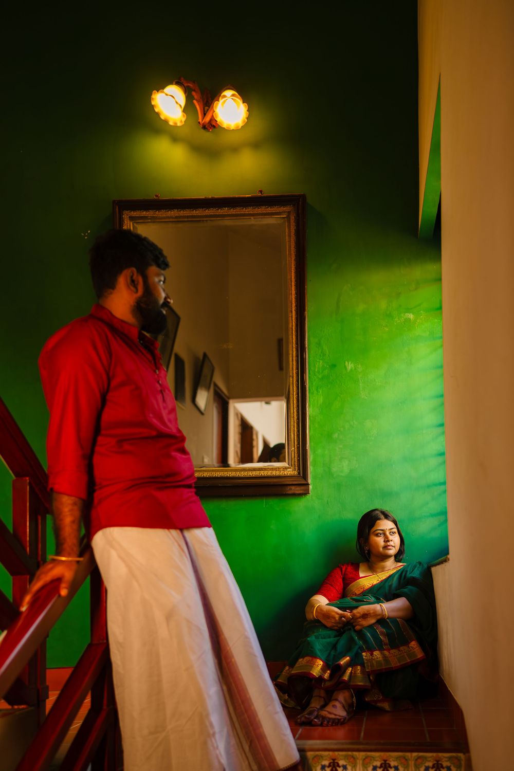 Couple in traditional wear pose in a green-walled room with dramatic lighting and mirror.