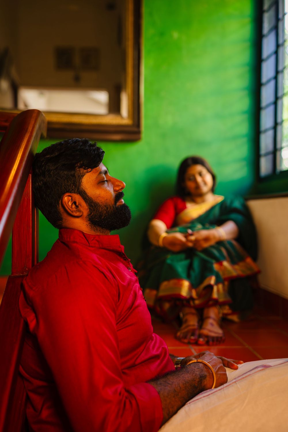 A couple in a room with green walls, one wearing red while the other sits relaxed on a couch.