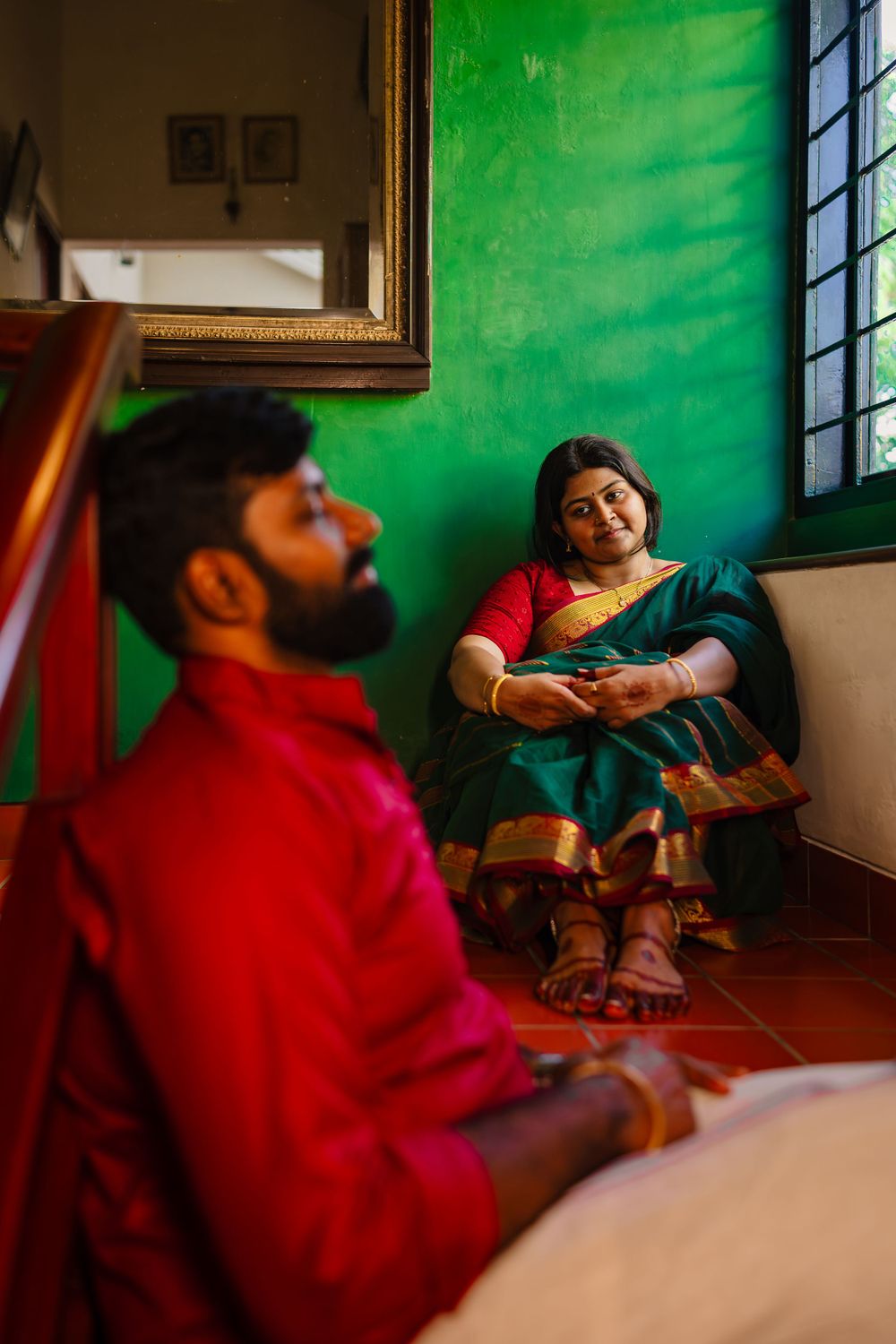 Two people in a room with green walls, one in red attire and another sitting casually.