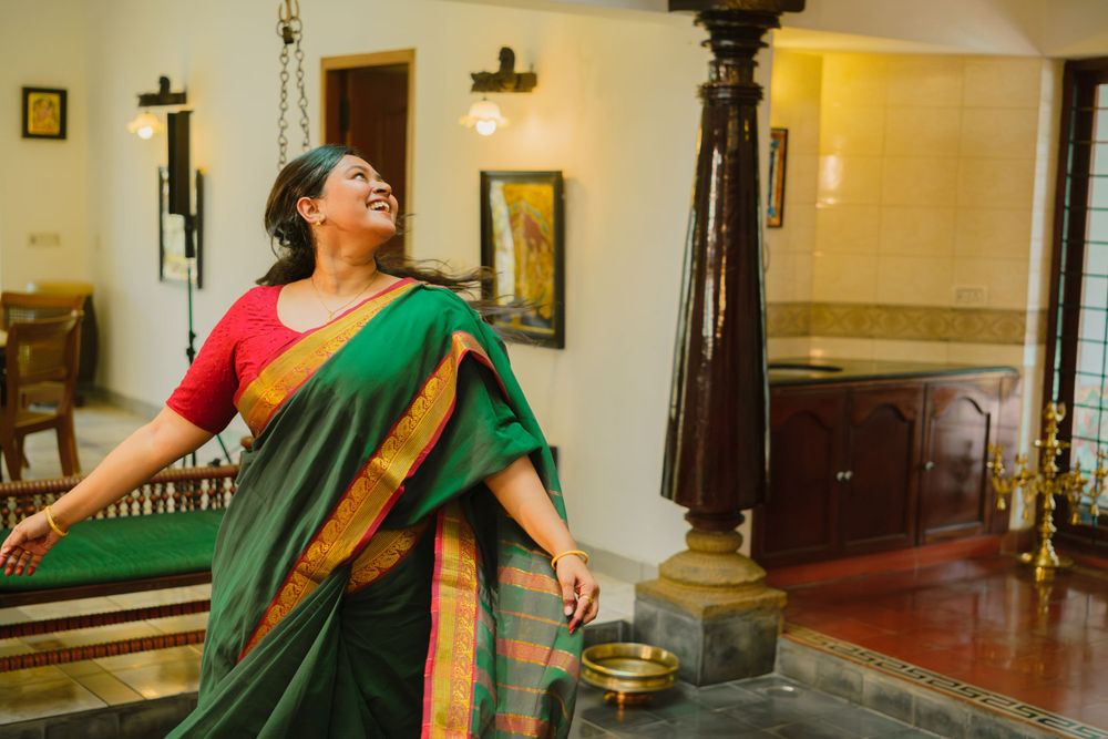 A person in a vibrant red and green sari strikes an expressive dance pose in a traditional interior setting with wooden pillars.